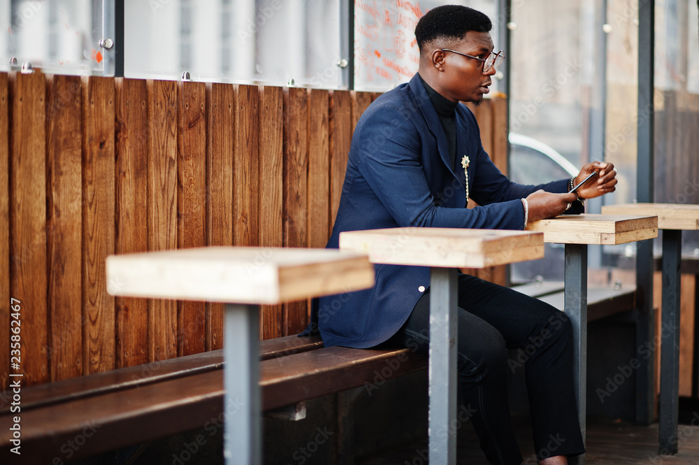 Amazingly looking african american man wear at blue blazer with brooch, black turtleneck and glasses posed at street. Fashionable black guy sitting behind table with mobile phone. Amazingly looking african american man wear at blue blazer with brooch, black turtleneck and glasses posed at street. Fashionable black guy sitting behind table with mobile phone.