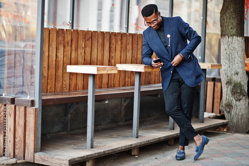Amazingly looking african american man wear at blue blazer with brooch, black turtleneck and glasses posed at street. Fashionable black guy with mobile phone. Amazingly looking african american man wear at blue blazer with brooch, black turtleneck and glasses posed at street. Fashionable black guy with mobile phone.