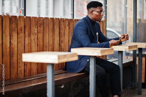 Amazingly looking african american man wear at blue blazer with brooch, black turtleneck and glasses posed at street. Fashionable black guy sitting behind table with mobile phone.