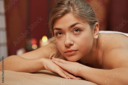 Healthy happy beautiful woman enjoying relaxing at spa center, looking away thoughtfully, copy space. Attractive woman lying on massage table, candles burning on background. Hotels, resorts concept