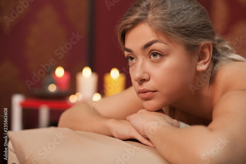 Young beautiful woman looking away dreamily, lying on massage table at spa center, candles on the back. Gorgeous blonde woman relaxing after spa treatment, copy space. Beauty, skincare, health concept