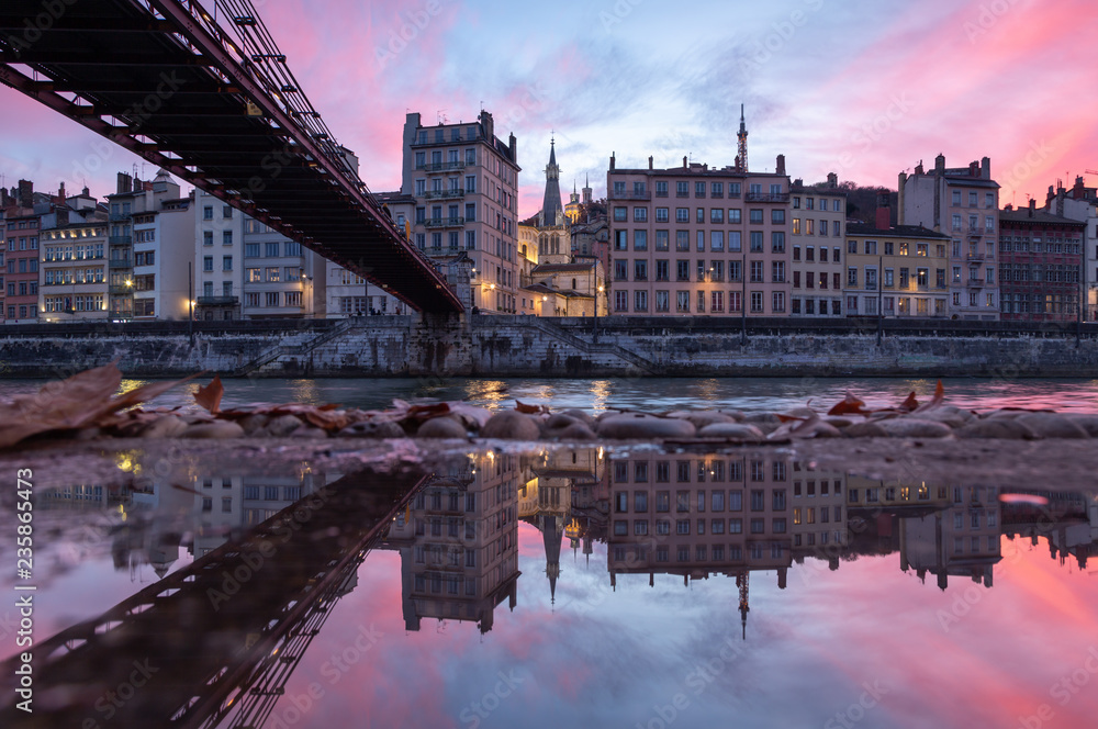 Old footbridge over the Saone river reflected in a puddle during a pink sunset in Vieux-Lyon, Lyon, France.