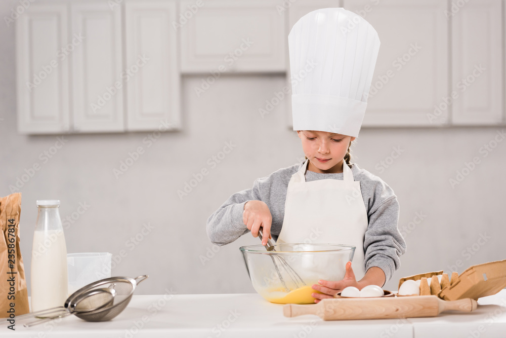 concentrated kid in chef hat whisking eggs in bowl at table in kitchen concentrated kid in chef hat whisking eggs in bowl at table in kitchen