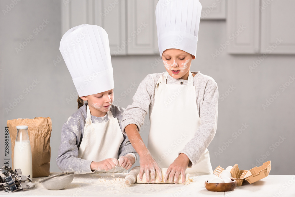 focused kids in aprons and chef hats making dough with rolling pin at table in kitchen focused kids in aprons and chef hats making dough with rolling pin at table in kitchen