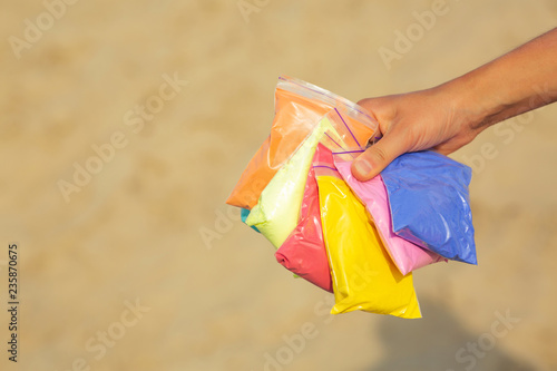 Closeup shot of female hand holding bags of dry colorful paint Holi against sand background. Empty space