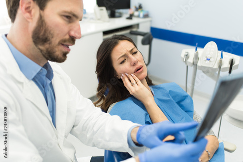 Young beautiful dentist is holding and looking at the teeth x-ray while woman patient is holding her hurtful tooth having painful face expression sitting at the modern dentist office.