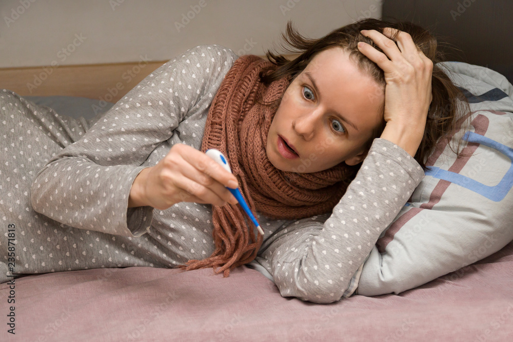 Young female is sick. She measuring temperature and looking at the thermometer in horror, lying in a bed at her home. High fever Young female is sick. She measuring temperature and looking at the thermometer in horror, lying in a bed at her home. High fever