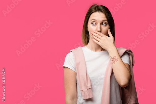 shocked young tattooed woman covering mouth by hand isolated on pink