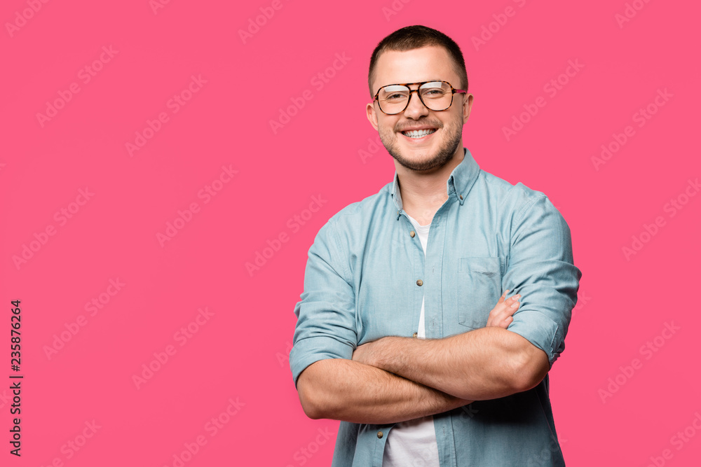 Fototapeta premium handsome happy man standing with crossed arms and smiling at camera isolated on pink