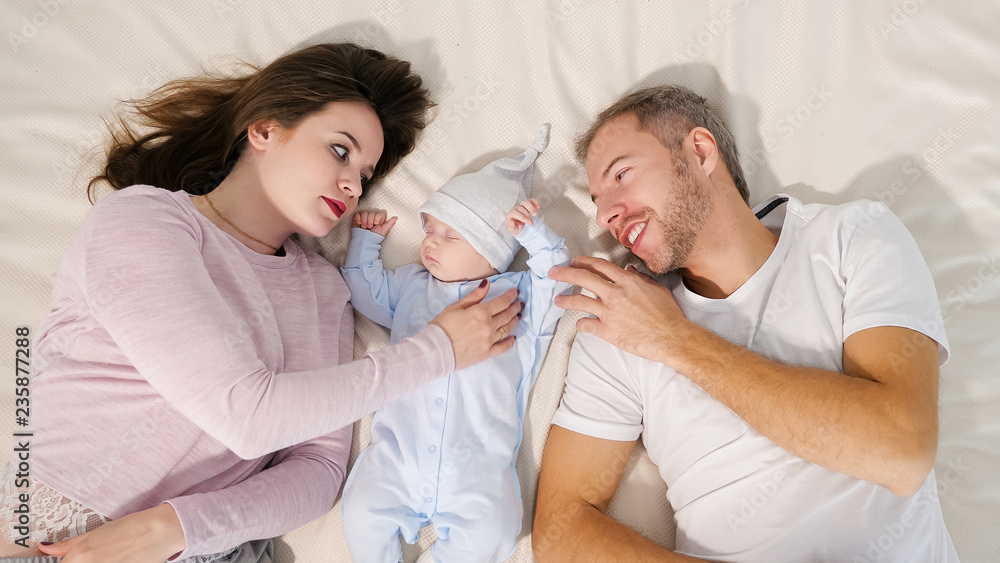 From above shot of happy mother and father lying on comfortable bed and looking at adorable sleeping boy
