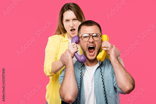 young woman holding handsets and man in eyeglasses yelling isolated on pink