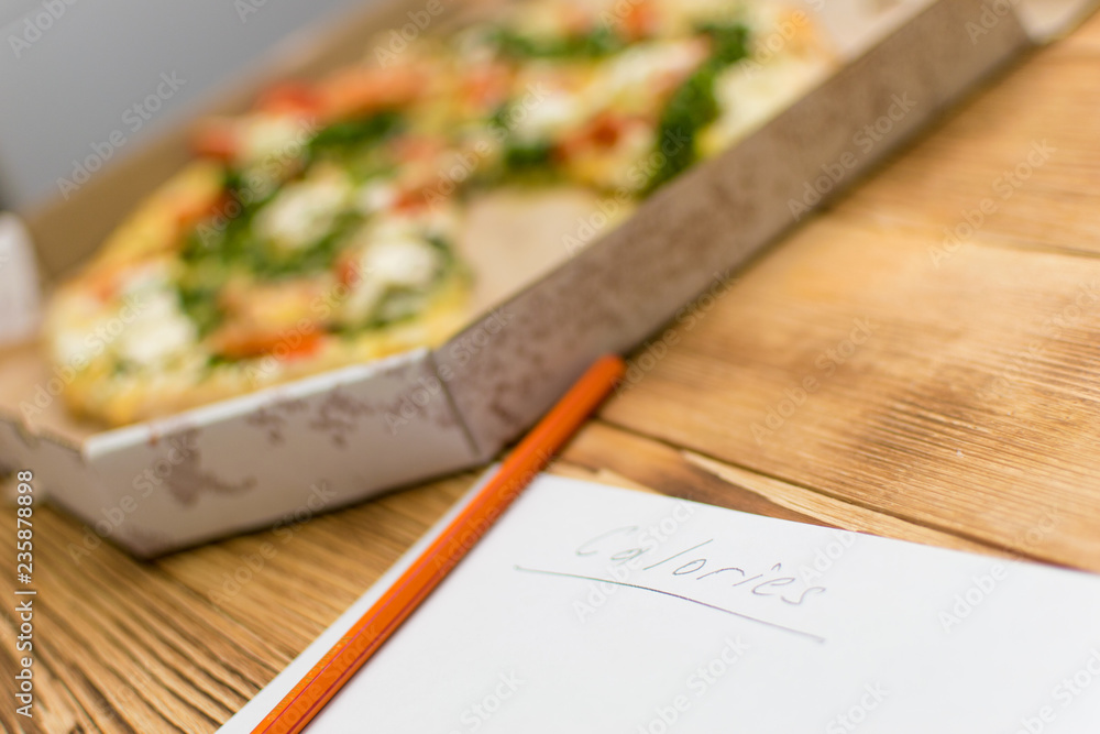 Count the calories. Sheet with the inscription Calories and lying near a pizza on a wooden background. Count the calories. Sheet with the inscription Calories and lying near a pizza on a wooden background.