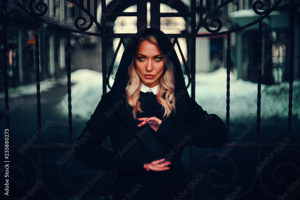 Fototapeta premium Mystical girl on the street in a mantle with a hood, on the background of an iron gate