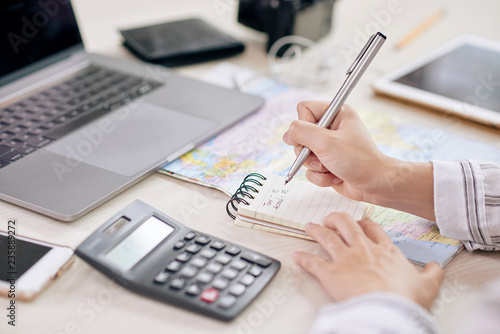 Crop shot of woman taking notes in notepad sitting at desk with laptop and calculating machine