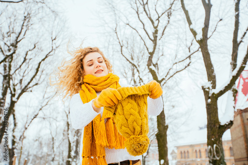 Outdoor close up portrait of blond curly playful hipster woman on street, looking at camera, smiling at snowy park. Model wearing white sweater, yelow winter hat, sarf, gloves. City lifestyle photo