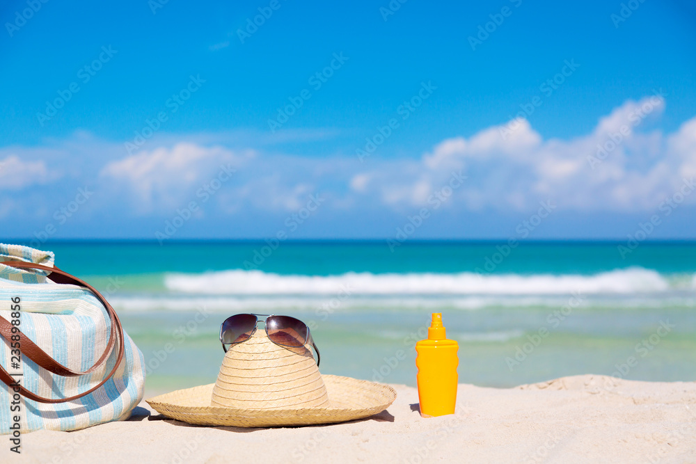 Beach accessories on sand for summer vacation concept. Bag, straw hat with sunglasses and sunscreen lotion bottle. White sand with amazing ocean and blue sky in the background.