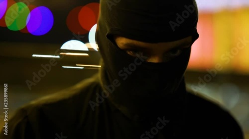 A man dressed in dark clothes and a balaclava goes with a bat in an abandoned park. There is an early winter outside and garlands hang around. His face is not visible in backlight. He is set up to fig photo