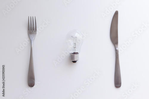 Light bulb  fork and knife on the white table. Electricity consumption  intellectual property concept. Top view  flat lay  copy space