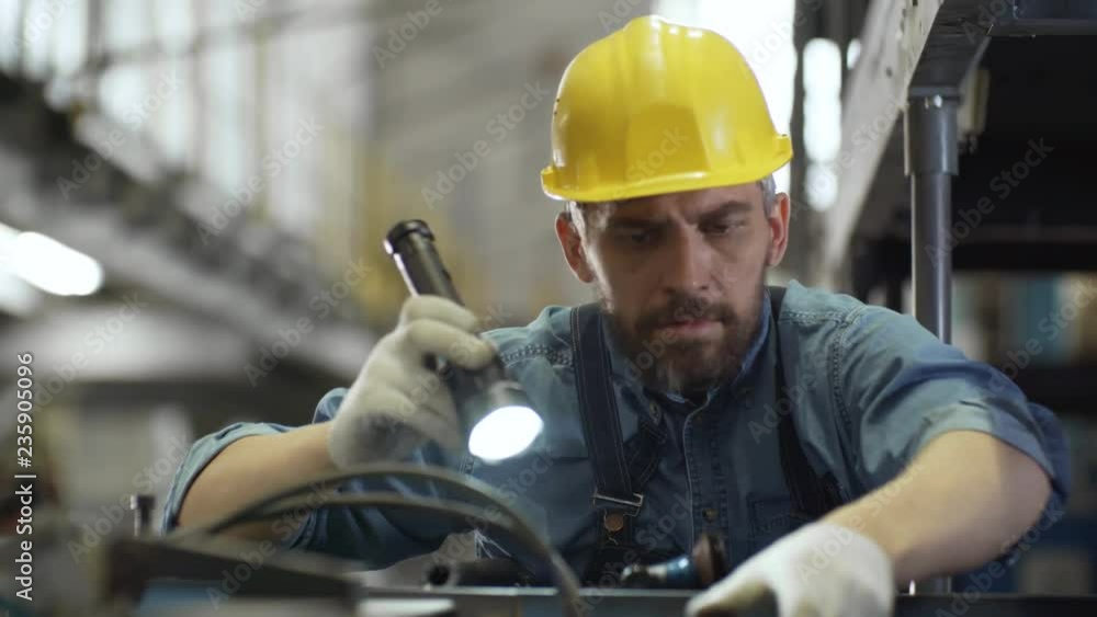 Tilt down of bearded Caucasian male technician in uniform and protective hardhat inspecting industrial equipment with flashlight while working in factory Tilt down of bearded Caucasian male technician in uniform and protective hardhat inspecting industrial equipment with flashlight while working in factory
