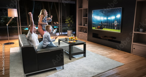A family is watching a soccer moment on the TV and celebrating a goal, sitting on the couch in the living room. © haizon