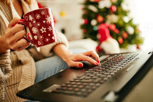 Woman holding cup of coffee while working on laptop at home for Christmas. New Year and Xmas concept.