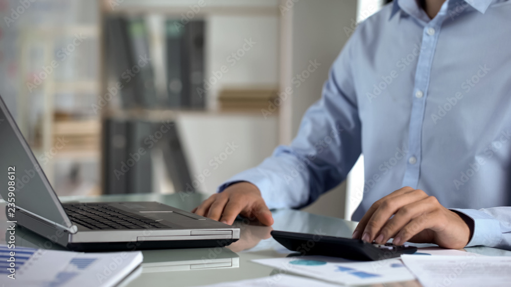 Office manager working on company sales report, using laptop and calculator Office manager working on company sales report, using laptop and calculator