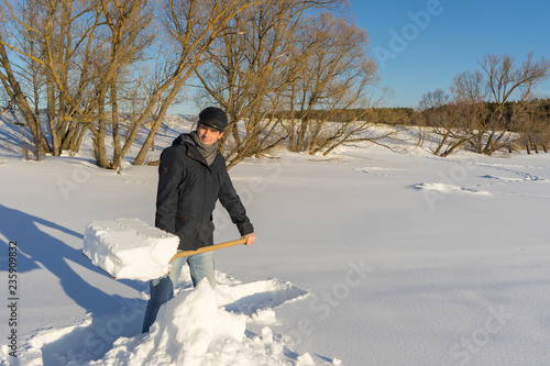 Handsome middle-aged caucasian man cleans snow on household plot in sunny day. Man with shovel paving path from country house to road. Winter active leisure concept