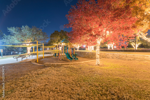 Holiday lightings at public park with playground near Dallas, Texas, USA. Christmas and New Year celebration. Beautiful Xmas decoration illumined string led on colorful fall foliage trees background