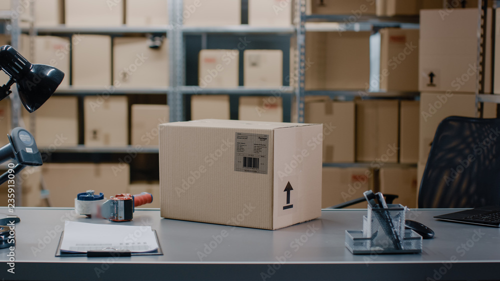 Cardboard Box Package Standing on the Table of the Warehouse where Rows of Shelves with Parcels Waiting to be Shipped and Delivered. Cardboard Box Package Standing on the Table of the Warehouse where Rows of Shelves with Parcels Waiting to be Shipped and Delivered.