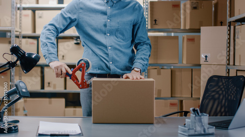 Professional Warehouse Worker Checks and Seales Cardboard Box Ready for Shipment. In the Background Person Working in the Rows of Shelves with Cardboard Boxes with Ready Orders. photo