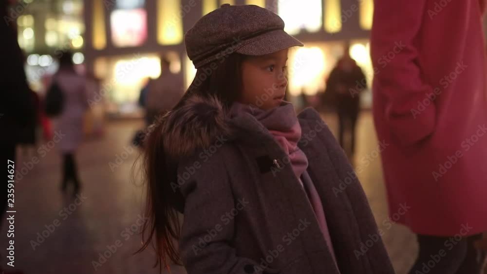 Child girl in a coat, cap and scarf is standing and waiting at the street against background of passing by people and evening city lights Child girl in a coat, cap and scarf is standing and waiting at the street against background of passing by people and evening city lights