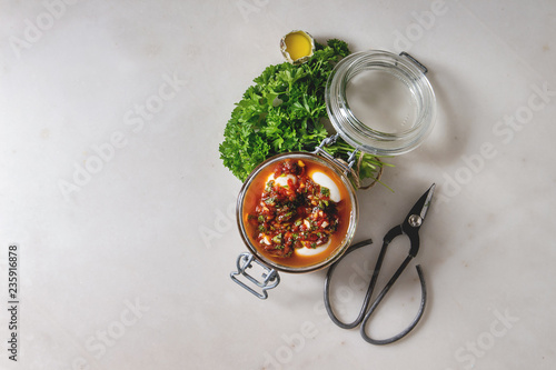Opened Jar with homemade pickled marinated quail eggs in tomato and olive oil sauce with anchovies and fresh parsley on white marble background. Ingredients above. Flat lay, space