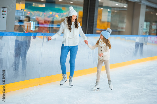 smiling mother and daughter holding hands and skating on ice rink together