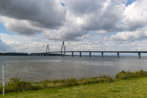 A blue sky with white fluffy cumulus clouds  a green meadow  and the Faro bridge with cable-stayed design that connect the islands of Falster and Zealand in Denmark.