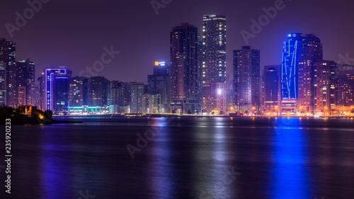 Beautiful view to Sharjah city center lights skyline at night, United Arab Emirates. Skyscrapers are reflecting in water