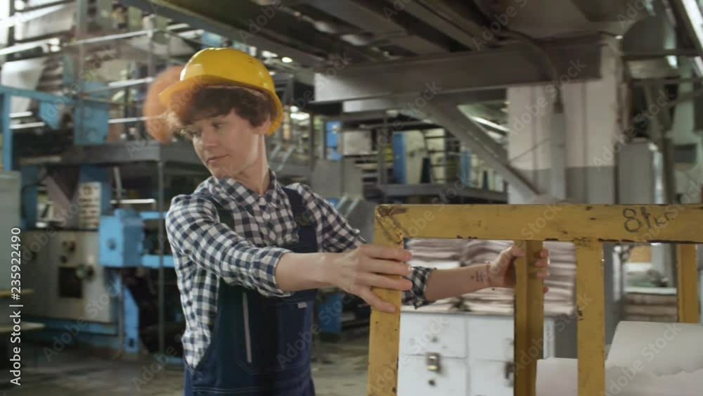 Young female worker in overall and hardhat pulling cart with paper reels while working in printing plant Young female worker in overall and hardhat pulling cart with paper reels while working in printing plant