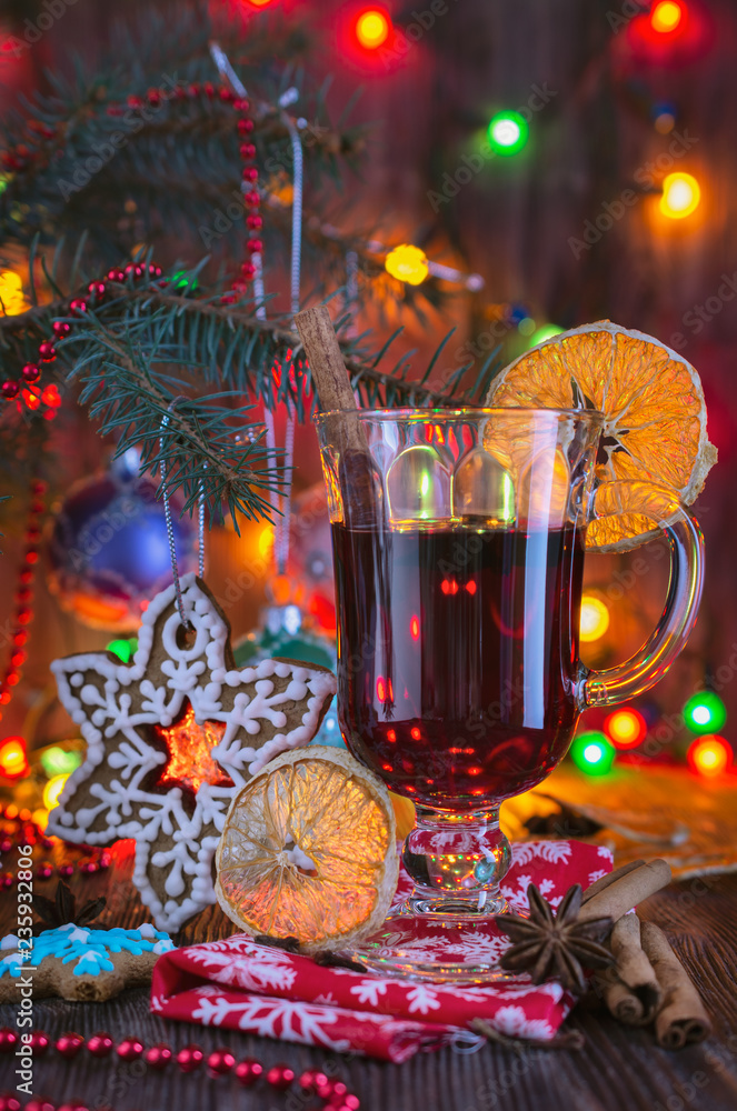 A glass of mulled wine with cinnamon and a slice of orange is on the table against the background of Christmas tree decorations on the branches of the Christmas tree in the light of Christmas lights i