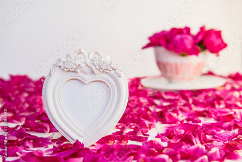 Heart shaped vintage style blank photoframe on the white table with fresh petals and bouquet of pink purple peonies in cup. Love, romantic, Valentines day concept. Selective focus. Copy space.