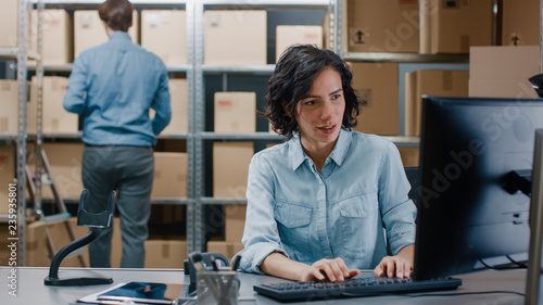 Female Inventory Manager Sitting at Her Desk and Using Personal Computer, Worker Puts Package on the Designated Shelf. In the Background Rows of Cardboard Boxes with Products Ready For Shipment. photo