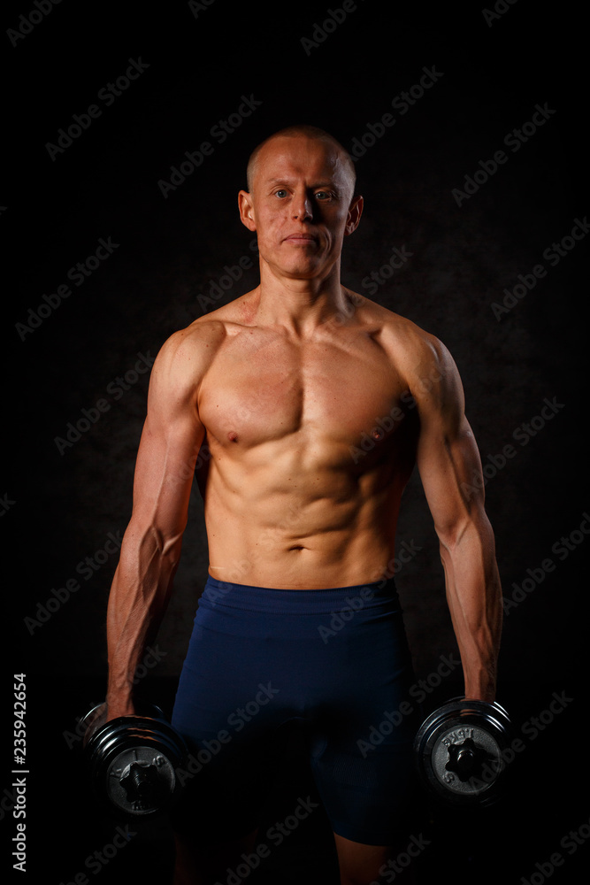 Handsome muscular man working out with dumbbells over black background.