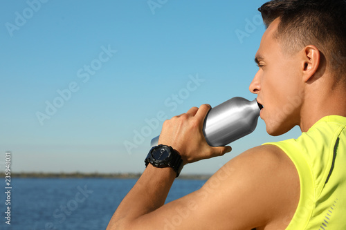 Young sporty man drinking from water bottle outdoors on sunny day. Space for text