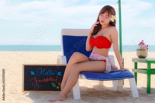 Sexy girl in red bikini sitting on chair beach under shadow of umbrella. Have a nice calligraphy board on side of chair. A lot sun and nice weather.