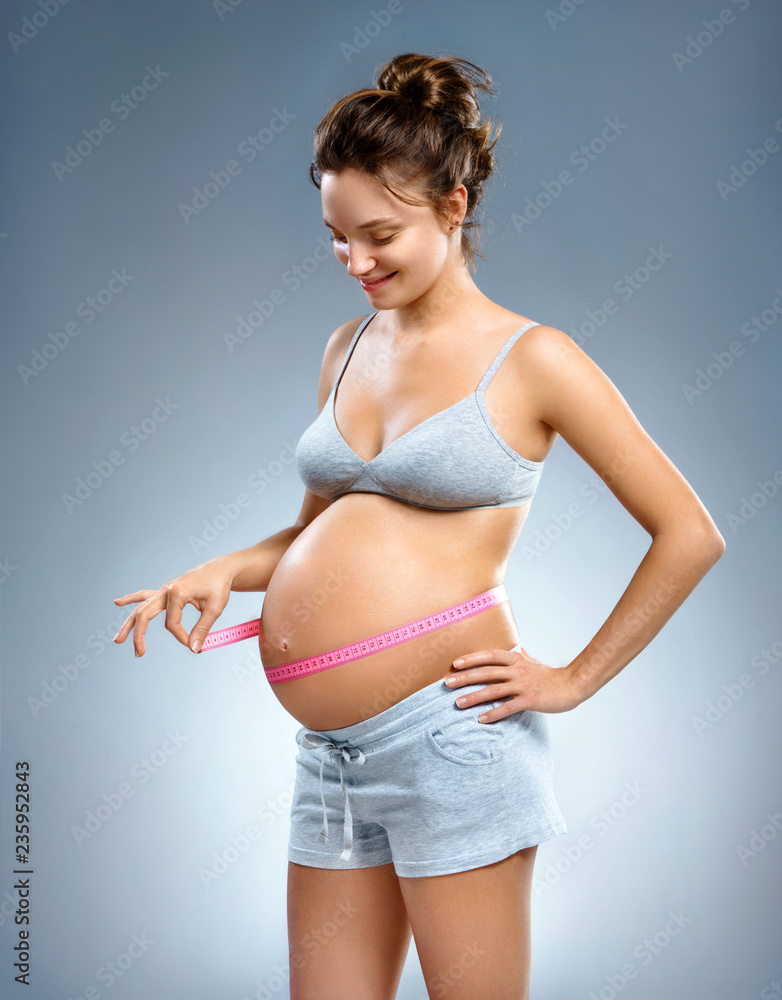 Obraz premium Smiling pregnant woman with measuring tape measures her belly on grey background. Pregnancy, maternity, preparation and expectation concept