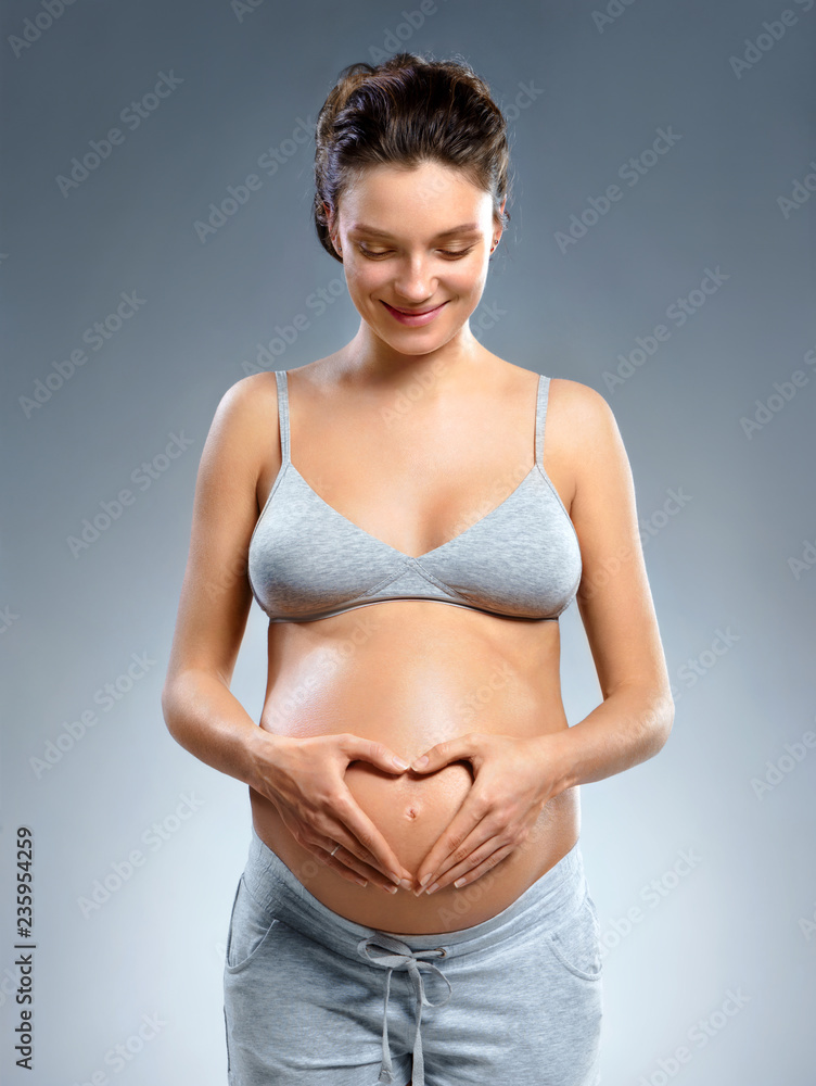 Smiling pregnant woman making a heart gesture with her fingers on grey background. Pregnancy, maternity, preparation and expectation concept Smiling pregnant woman making a heart gesture with her fingers on grey background. Pregnancy, maternity, preparation and expectation concept