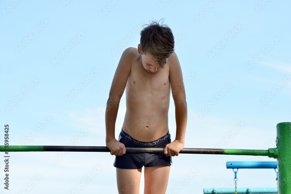A boy in shorts goes in for sports on a horizontal bar on the street. Does  various exercises, pull-ups, pushups and hangs upside down. Photos | Adobe  Stock