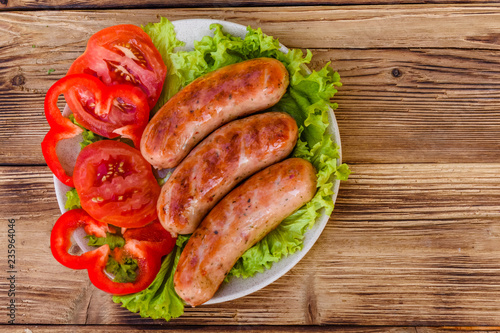 Ceramic plate with grilled sausages, sliced tomatoes and lettuce leaves on wooden table. Top view