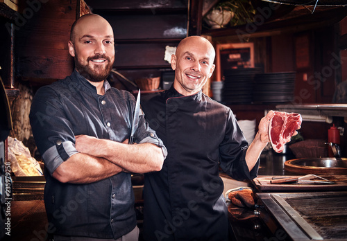 Chef holding a fresh steak and his assistant standing near in a restaurant kitchen.