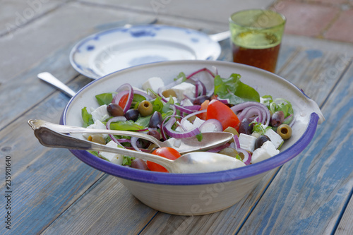 Bowl filled with sallad, tomato, red onion, olives and sheep cheese. An old fashioned white and blue plate, forke, knife and a glass of beer on a blue wethered wooden table