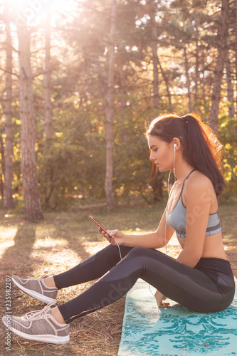 Sports lifestyle. young and beautiful athletic girl in a sports top and leggings sits on a mat after a workout with headphones and a telephone in her hands in the sunset rays in a park. vertical photo