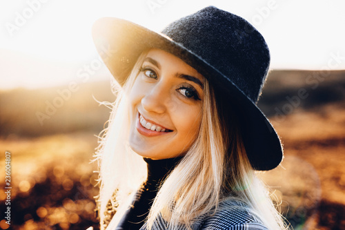 Pretty young woman in stylish hat cheerfully smiling and looking at camera while standing on blurred background of autumn nature on sunny day.Cheerful female in countryside